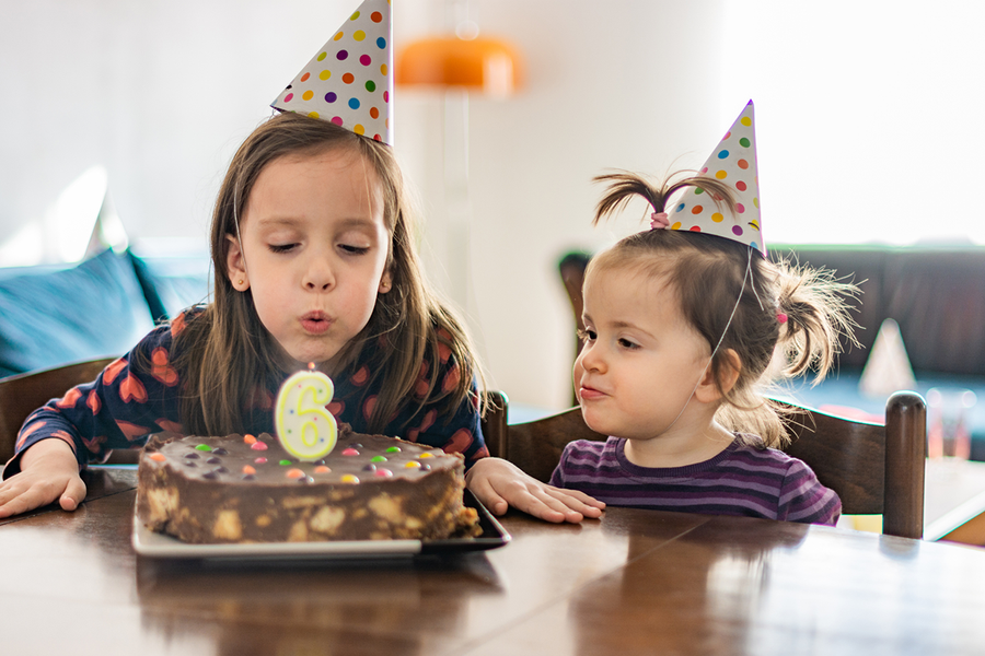 A young girl in a party hat blowing out a candle on her 6th birthday cake with a friend, showing birthday gift ideas and birthday presents for children from Photobox.