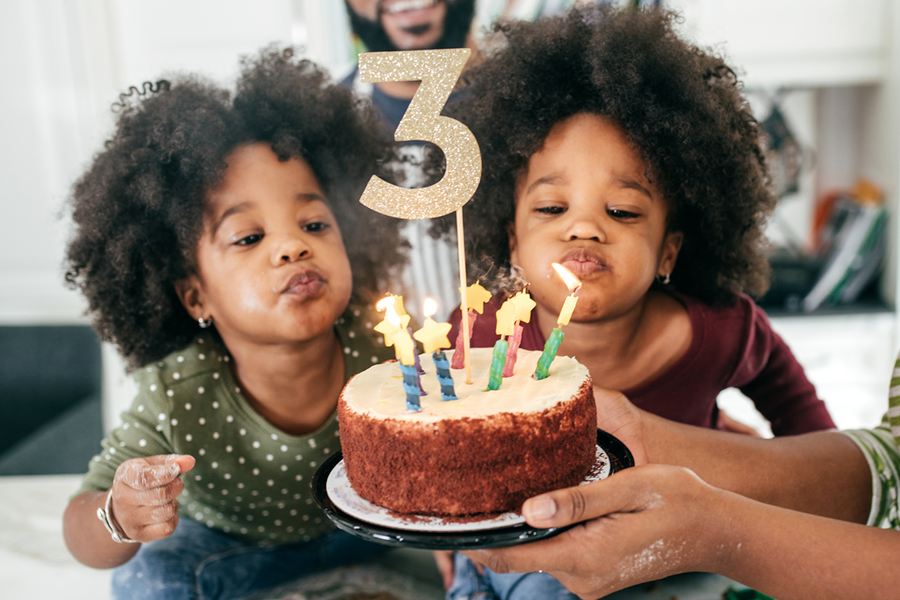 Two twin girls about to blow out the candles on their 3rd birthday cake, showing birthday presents for her and personalised birthday gifts from Photobox.
