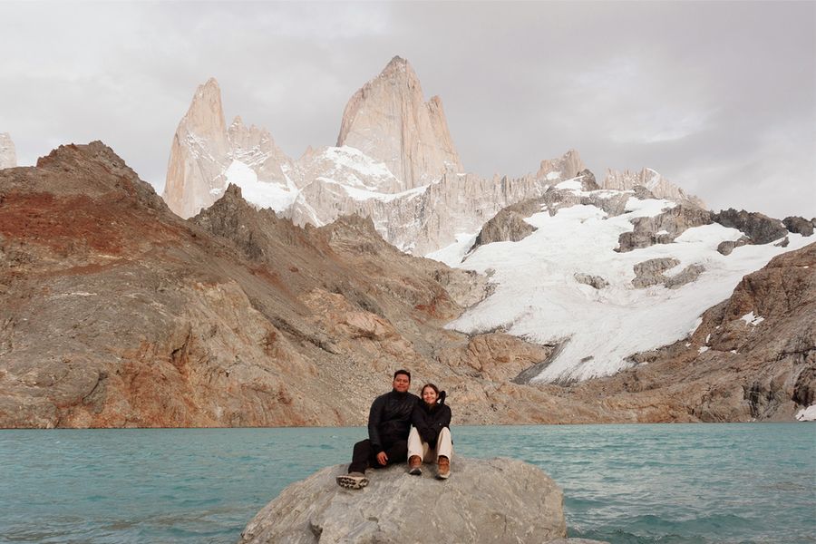 A couple sitting on a rock in front of a body of water and snowy mountains, showcasing an extreme long shot and landscape framing techniques with Photobox.