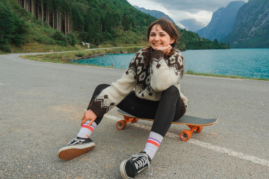 A woman smiling while sitting on a skateboard in front of a lake and mountains, demonstrating a full shot and how to frame subjects in photography with Photobox.