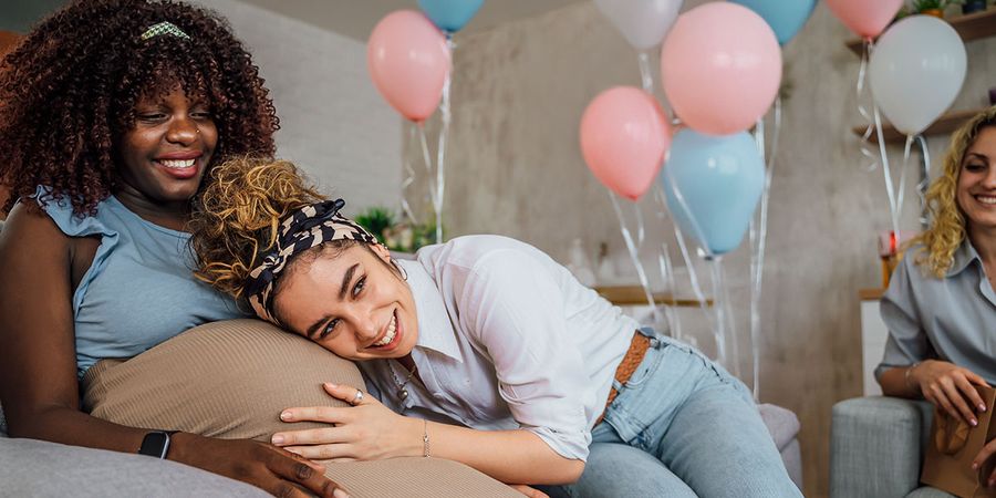 A pregnant woman at her baby shower with a friend listening to her belly, highlighting baby shower ideas and baby shower decorations from Photobox.