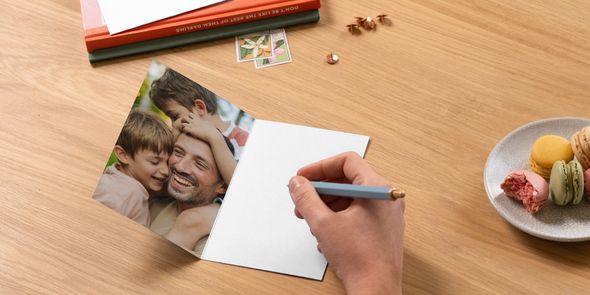 An open personalised Photobox Father’s Day photo card shows a father with his two sons, with a pen poised to write a message beside books and a plate of macarons, inspiring what to write in a Father’s Day card.