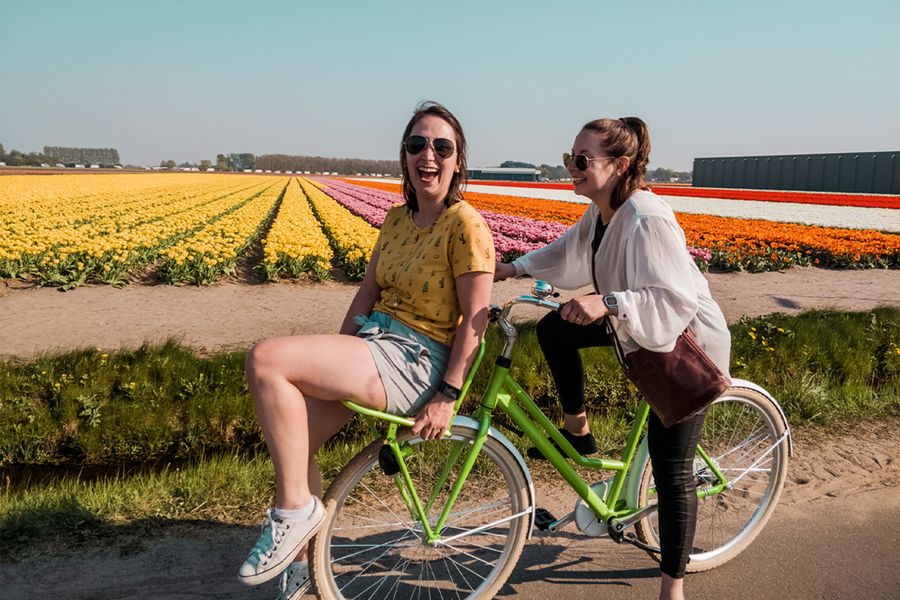 Two women having fun on a bike beside a colourful flower field, inspiring Photobox spring photo ideas and spring photography that captures flowers, colour and the joy of the season.