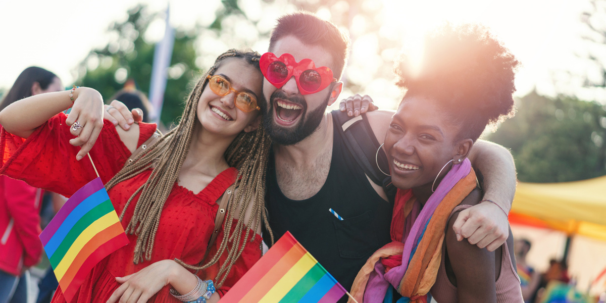 Three friends smiling at a Pride event, holding rainbow flags and wearing sunglasses, celebrating the joy and unity of LGBTQI+ Pride.