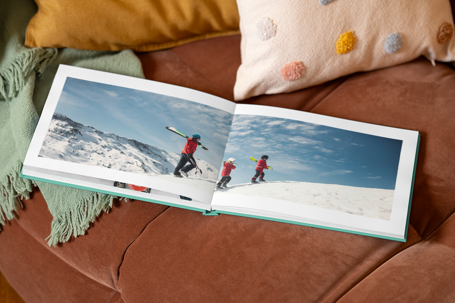 An open premium layflat photo book from Photobox resting on a sofa, displaying a stunning full-page spread of a snowy holiday photo, perfect for a personalised photo album of your travels.