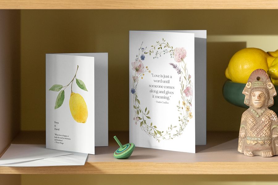 Two Photobox personalised wedding invitations with floral and lemon designs displayed on a shelf beside a fruit bowl and ornament, inspiring wedding card messages and personal wedding invitation wording.