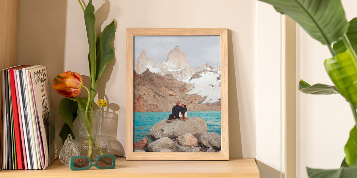 A framed landscape photo leaning on a wooden shelf next to books and flowers in a vase, showing how to display wall art without nails using Photobox.