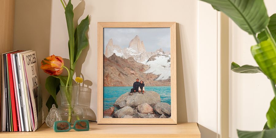A framed landscape photo leaning on a wooden shelf next to books and flowers in a vase, showing how to display wall art without nails using Photobox.