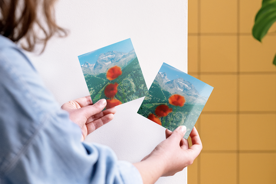A woman comparing two Photobox photo prints of the same mountain landscape with flowers in focus, showing matte photo prints and gloss photo prints to help choose the right finish for photo printing.
