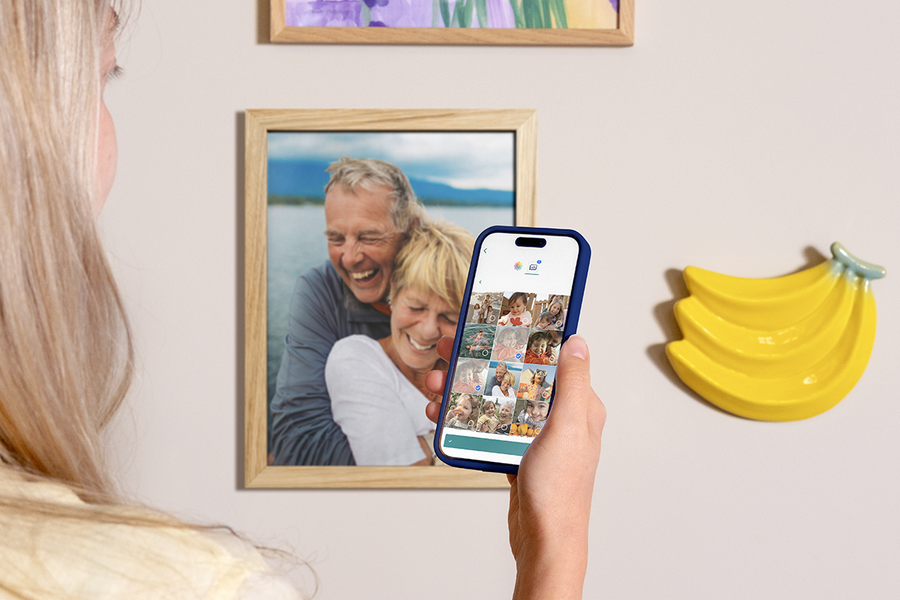 A woman holding up her phone to show how easy it is to order Photobox photo prints from the app, with framed photo prints displayed on the wall behind her, inspiring photo printing online and printing photos from your phone.