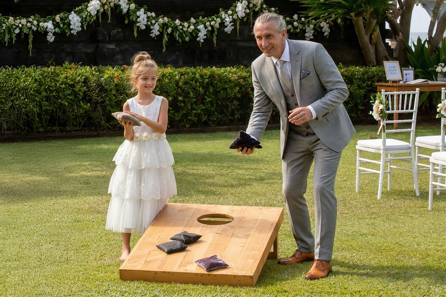 A young girl in formal wedding attire and an older man in a suit playing wedding cornhole, throwing bean bags into the board, inspiring Photobox wedding games and outdoor wedding games for guests.